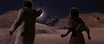 Movie still from “Tobruk” (1967), directed by Arthur Hiller – Two people are standing in the sand and one is holding up a gun; Wide shot, Over the shoulder angle