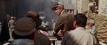 Movie still from “Tobruk” (1967), directed by Arthur Hiller – A man wearing a hat sitting on top of a railing next to a crowd; Medium shot, Over the shoulder angle