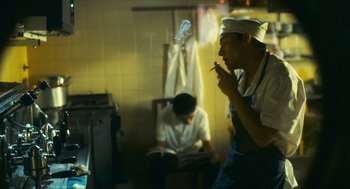 Movie still from “Happy Together” (1997), directed by Kar-Wai Wong – A man smoking a cigarette while another man reads a book in the background; Medium shot, Low angle