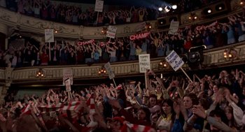 Movie still from “Tommy” (1975), directed by Ken Russell – A group of people holding signs in the air in an auditorium; Extreme Wide shot, High angle