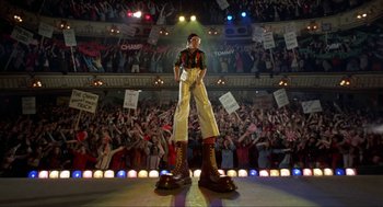 Movie still from “Tommy” (1975), directed by Ken Russell – A man standing on top of a stage in front of an audience; Wide shot, Low angle
