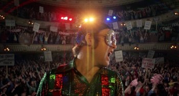Movie still from “Tommy” (1975), directed by Ken Russell – A man in a sequined shirt and glasses is in front of an audience; Medium shot, Low angle