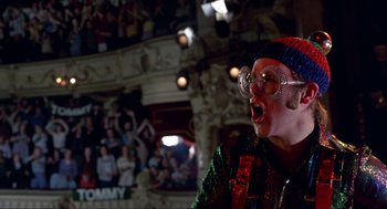 Movie still from “Tommy” (1975), directed by Ken Russell – A person wearing a clown wig and glasses; Medium shot, Low angle