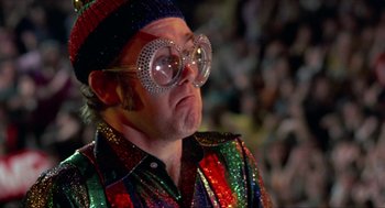 Movie still from “Tommy” (1975), directed by Ken Russell – A man wearing a pair of glasses and a colorful shirt; Close Up shot, Low angle