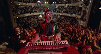 Movie still from “Tommy” (1975), directed by Ken Russell – A man in a clown costume playing a keyboard; Wide shot, Overhead angle