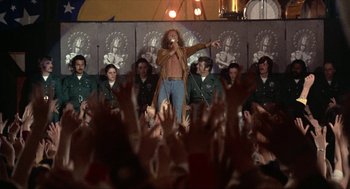Movie still from “Tommy” (1975), directed by Ken Russell – A group of people are sitting in front of a crowd; Wide shot, Low angle