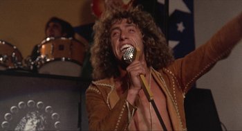 Movie still from “Tommy” (1975), directed by Ken Russell – A man in a brown jacket is singing into a microphone; Medium shot, Low angle