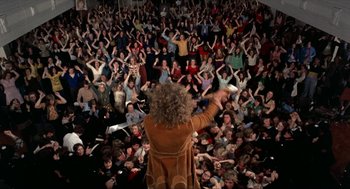 Movie still from “Tommy” (1975), directed by Ken Russell – A crowd of people in a room with a person holding a microphone in their hand; Wide shot, Overhead angle