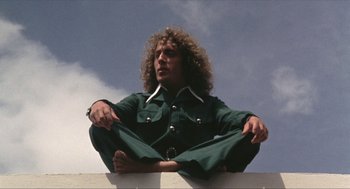 Movie still from “Tommy” (1975), directed by Ken Russell – A man sitting on top of a building with his legs crossed; Medium shot, Low angle