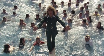 Movie still from “Tommy” (1975), directed by Ken Russell – A man standing on a surfboard in the middle of the water; Wide shot, High angle
