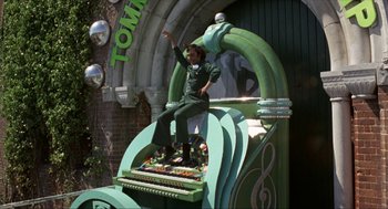 Movie still from “Tommy” (1975), directed by Ken Russell – A man sitting on top of an organ in front of a building; Wide shot, Low angle