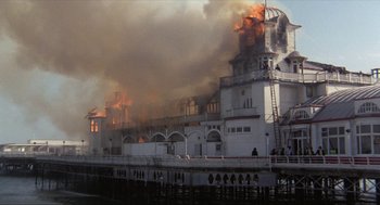 Movie still from “Tommy” (1975), directed by Ken Russell – A large building on fire on the side of a pier; Extreme Wide shot, Low angle