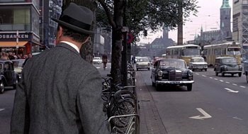 Movie still from “Topaz” (1969), directed by Alfred Hitchcock – A man wearing a suit and a hat standing next to a tree; Wide shot, Over the shoulder angle