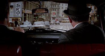 Movie still from “Topaz” (1969), directed by Alfred Hitchcock – A man in a hat driving a car down a city street; Wide shot, Over the shoulder angle