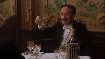 Movie still from “Topsy-Turvy” (1999), directed by Mike Leigh – A man sitting at a dinner table holding a wine glass; Medium shot, Low angle