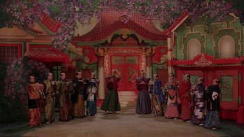 Movie still from “Topsy-Turvy” (1999), directed by Mike Leigh – A group of people dressed in traditional asian garb in front of a building; Wide shot, High angle