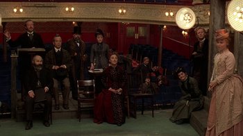 Movie still from “Topsy-Turvy” (1999), directed by Mike Leigh – A group of people sitting and standing on a stage; Wide shot, High angle