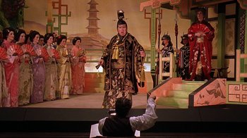 Movie still from “Topsy-Turvy” (1999), directed by Mike Leigh – A man sitting in front of an audience holding a sword; Wide shot, Low angle