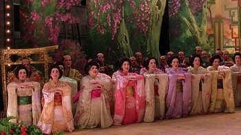 Movie still from “Topsy-Turvy” (1999), directed by Mike Leigh – A group of people dressed in geishas sitting in front of a tree; Wide shot, High angle