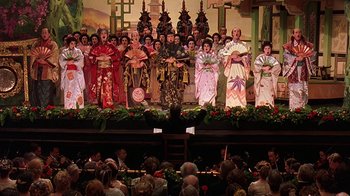 Movie still from “Topsy-Turvy” (1999), directed by Mike Leigh – A group of people in traditional asian garb on a stage; Extreme Wide shot, High angle