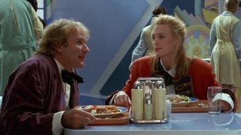 Movie still from “Toys” (1992), directed by Barry Levinson – A man and a woman sitting at a table with plates of food in front of them; Medium shot, Over the shoulder angle