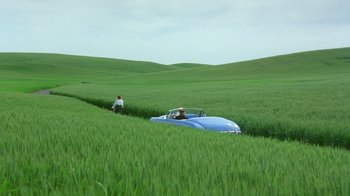 Movie still from “Toys” (1992), directed by Barry Levinson – A man standing next to a blue car in the middle of a green field; Extreme Wide shot, High angle