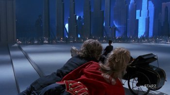 Movie still from “Toys” (1992), directed by Barry Levinson – A couple of people that are sitting in front of some buildings; Extreme Wide shot, High angle