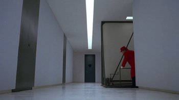 Movie still from “Toys” (1992), directed by Barry Levinson – A man in a red suit and a red hat is standing in front of a door; Wide shot, High angle