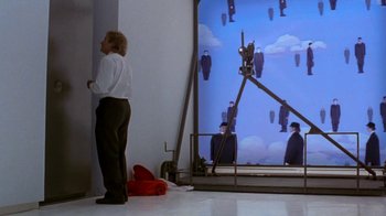 Movie still from “Toys” (1992), directed by Barry Levinson – A man standing in front of an image of people in suits; Wide shot, Low angle