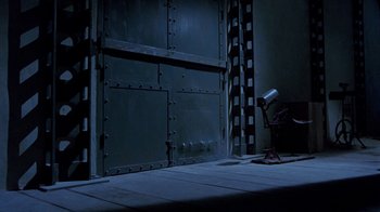 Movie still from “Toys” (1992), directed by Barry Levinson – A dark room with a metal door and a light; Wide shot, High angle