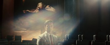 Movie still from “Trance” (2013), directed by Danny Boyle – A man in a white suit and red neck tie sitting in front of a projection screen; Medium shot, Low angle