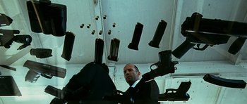 Movie still from “Transporter 3” (2008), directed by Olivier Megaton – A man in a suit and tie holding a gun in front of a bunch of guns; Medium shot, Overhead angle