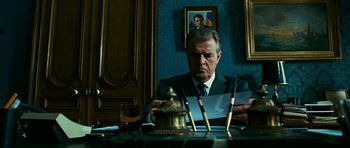 Movie still from “Transporter 3” (2008), directed by Olivier Megaton – A man sitting at a table in front of a painting; Medium shot, Low angle