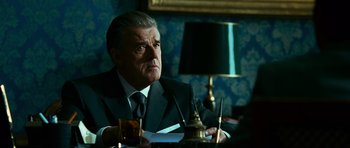 Movie still from “Transporter 3” (2008), directed by Olivier Megaton – An older man sitting at a table in front of a lamp; Close Up shot, Low angle