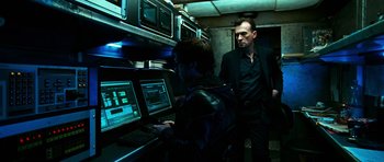 Movie still from “Transporter 3” (2008), directed by Olivier Megaton – A man standing next to another man using a laptop computer; Medium shot, Low angle