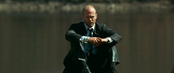 Movie still from “Transporter 3” (2008), directed by Olivier Megaton – A man in a suit and tie sitting on the ground looking at his watch; Medium shot, Low angle