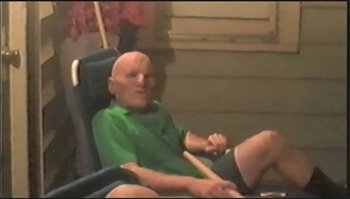 Movie still from “Trash Humpers” (2009), directed by Harmony Korine – An older man sitting in a chair holding a drum stick; Medium shot, High angle