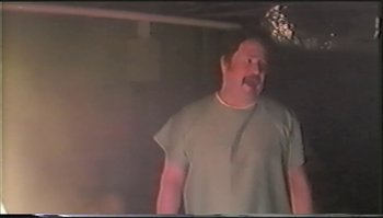 Movie still from “Trash Humpers” (2009), directed by Harmony Korine – A man standing in a room wearing a t - shirt; Medium shot, Low angle