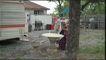 Movie still from “Trash Humpers” (2009), directed by Harmony Korine – An older woman sitting at an outdoor table in a yard; Wide shot, High angle