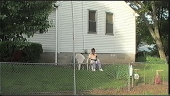 Movie still from “Trash Humpers” (2009), directed by Harmony Korine – A man sitting on a lawn chair in a yard next to a fence; Extreme Wide shot, High angle