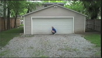 Movie still from “Trash Humpers” (2009), directed by Harmony Korine – A person crouching down in front of a garage door; Extreme Wide shot, High angle