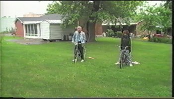 Movie still from “Trash Humpers” (2009), directed by Harmony Korine – Two people riding bikes in a grassy yard; Extreme Wide shot, High angle