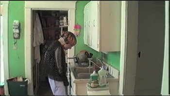 Movie still from “Trash Humpers” (2009), directed by Harmony Korine – A person standing in a kitchen next to a sink; Medium shot, High angle
