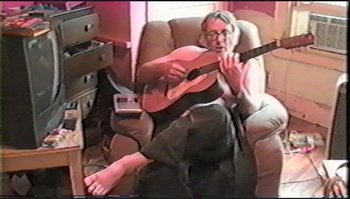 Movie still from “Trash Humpers” (2009), directed by Harmony Korine – A man sitting in a chair playing a guitar; Medium shot, High angle