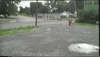 Movie still from “Trash Humpers” (2009), directed by Harmony Korine – A person riding a skate board down a street; Extreme Wide shot, High angle