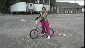 Movie still from “Trash Humpers” (2009), directed by Harmony Korine – A woman standing next to a bicycle in a parking lot; Wide shot, High angle