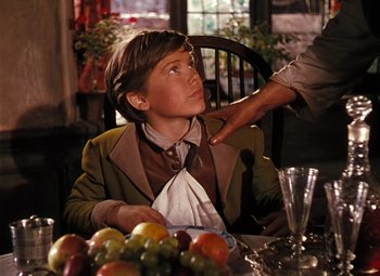 Movie still from “Treasure Island” (1950), directed by Byron Haskin – A young boy sitting at a dinner table with a man touching his ear; Medium shot, Over the shoulder angle