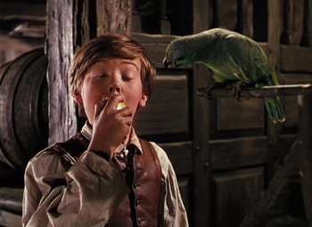 Movie still from “Treasure Island” (1950), directed by Byron Haskin – A young boy eating a piece of food while a parrot sits on a tree; Medium shot, Over the shoulder angle