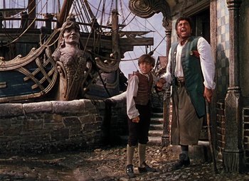 Movie still from “Treasure Island” (1950), directed by Byron Haskin – A man standing next to a young boy in front of a building; Wide shot, Low angle