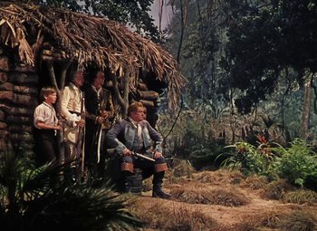 Movie still from “Treasure Island” (1950), directed by Byron Haskin – A group of people standing in front of a hut in the woods; Wide shot, Low angle